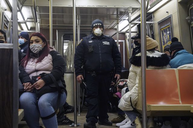 A masked police officer wearing an NYPD knit cap and a jacket that says NYPD stands in the middle of a train; other passenger are seated.